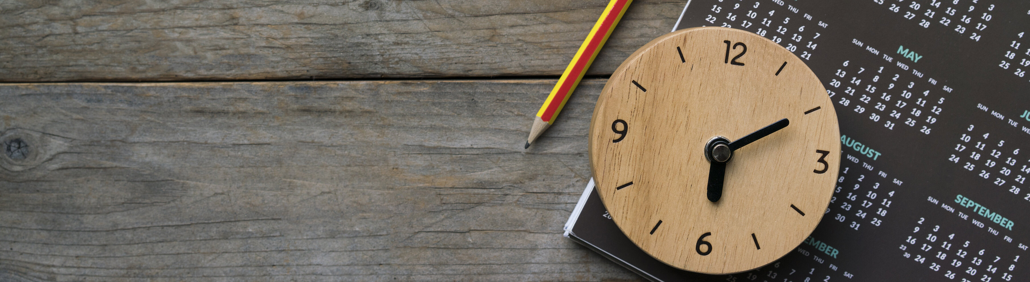 Auf einem Holztisch liegt ein Kalender neben dem ein Bleistift liegt und eine kleine Holzuhr