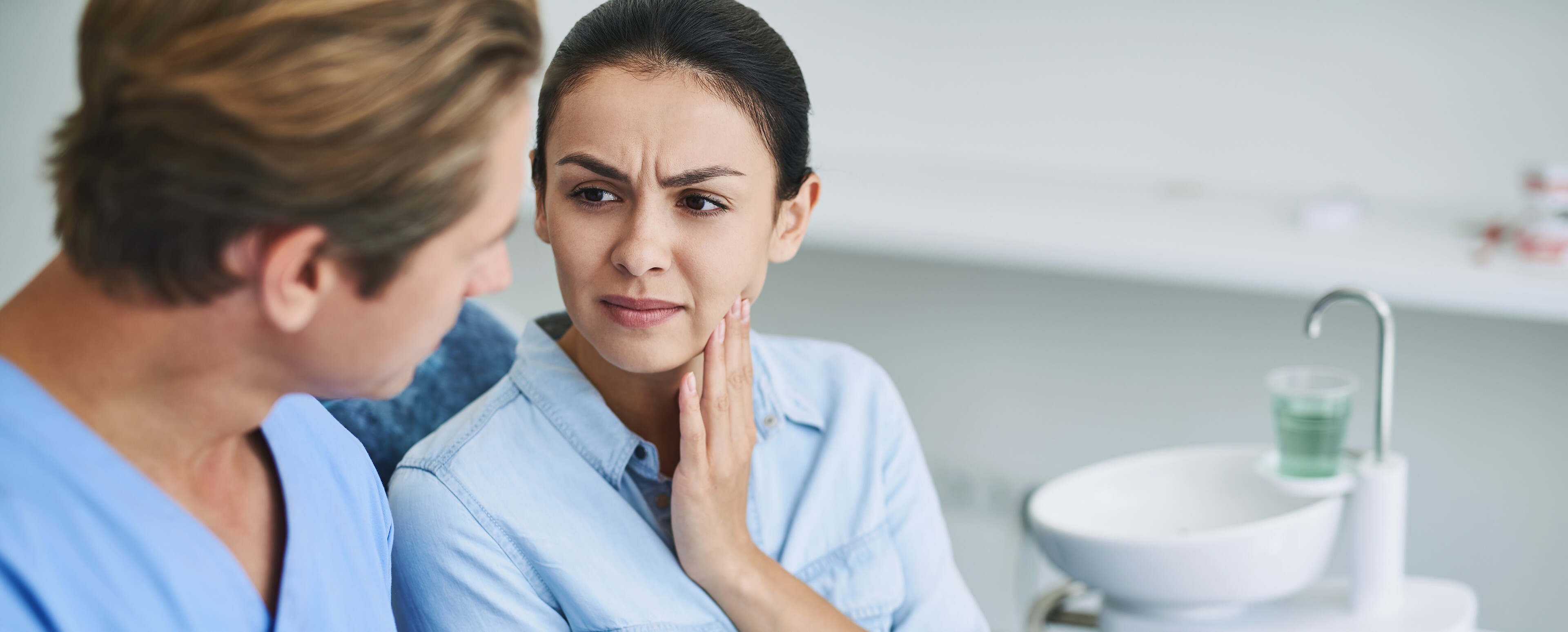 Patientin hält sich wegen Kieferschmerzen mit einer Hand die Wange während sie von einem Zahnarzt beraten wird