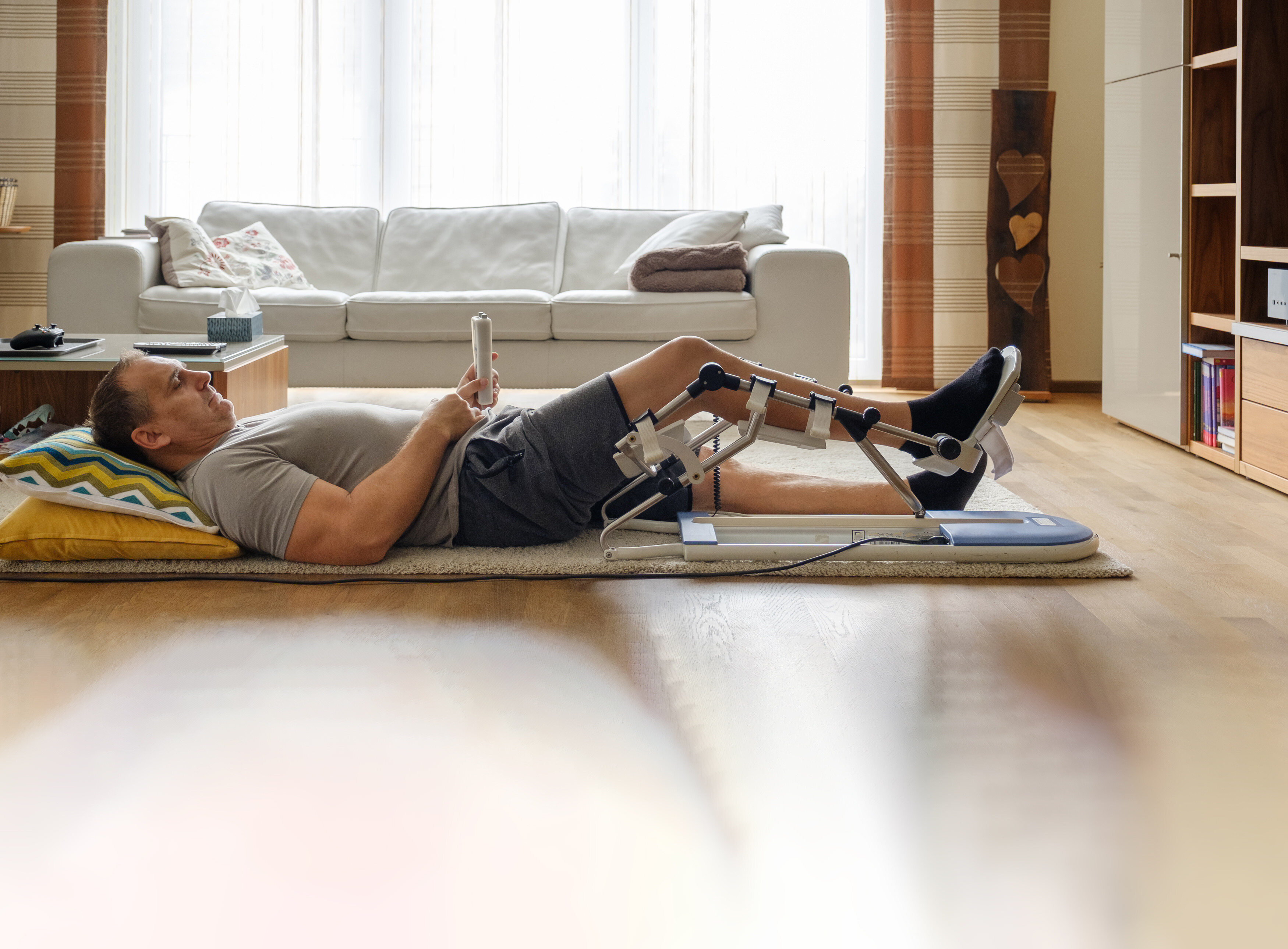 Mann liegt entspannt mit einer CPM Beinmotorschiene am rechten Bein auf dem Rücken auf einem Wohnzimmerteppich