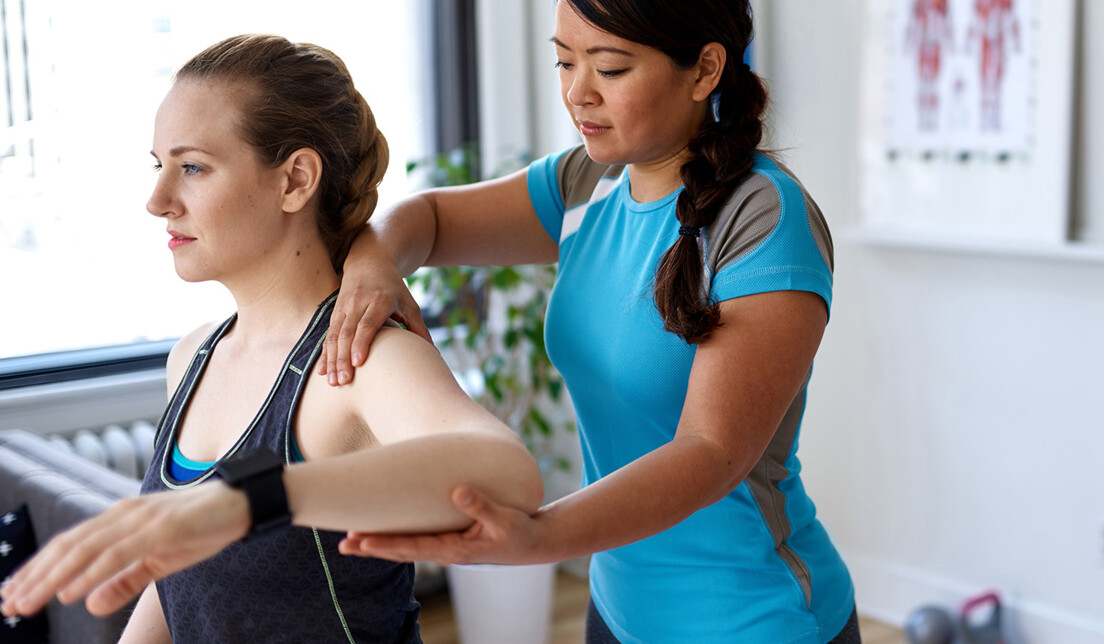 Eine Physiotherapeutin steht hinter einer Patientin und hilft dieser bei einer Schulterübung